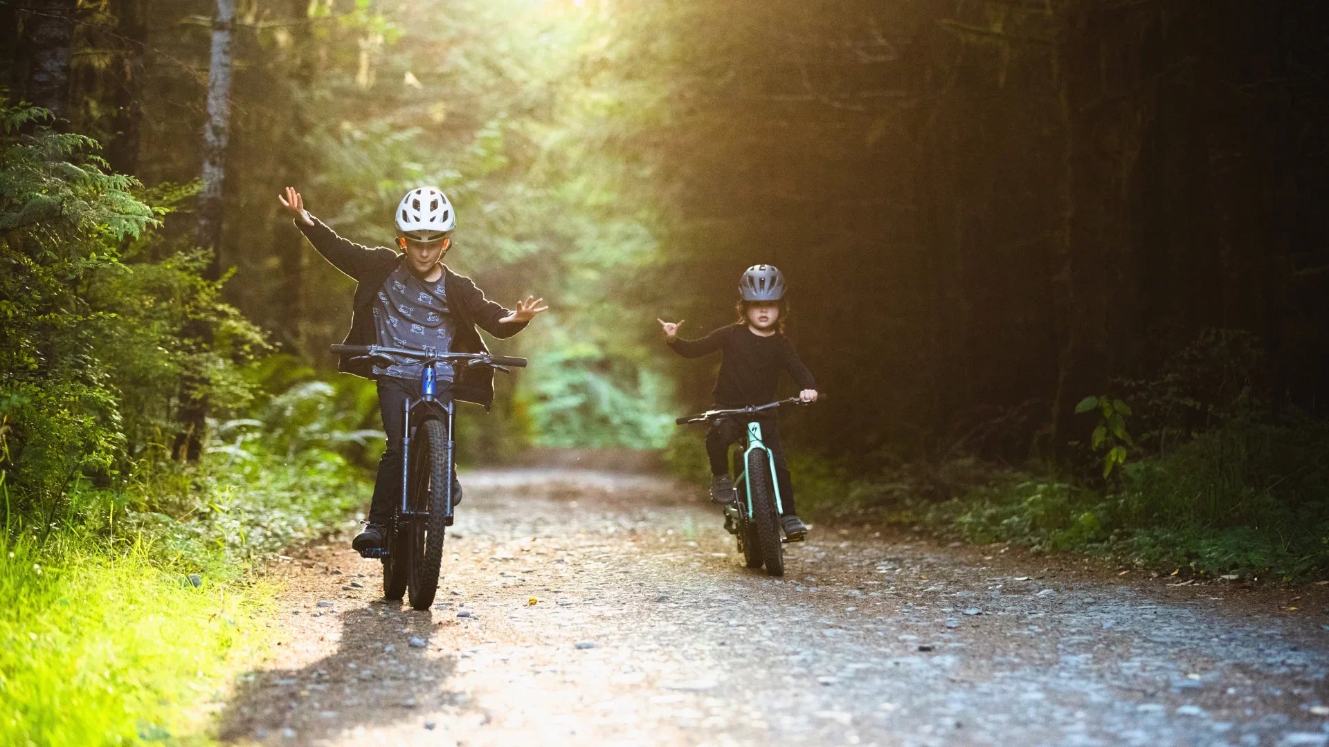 Two children ride bikes on a sunlit forest trail, both wearing helmets and smiling, with one child lifting both hands off the handlebars while balancing on the bike.