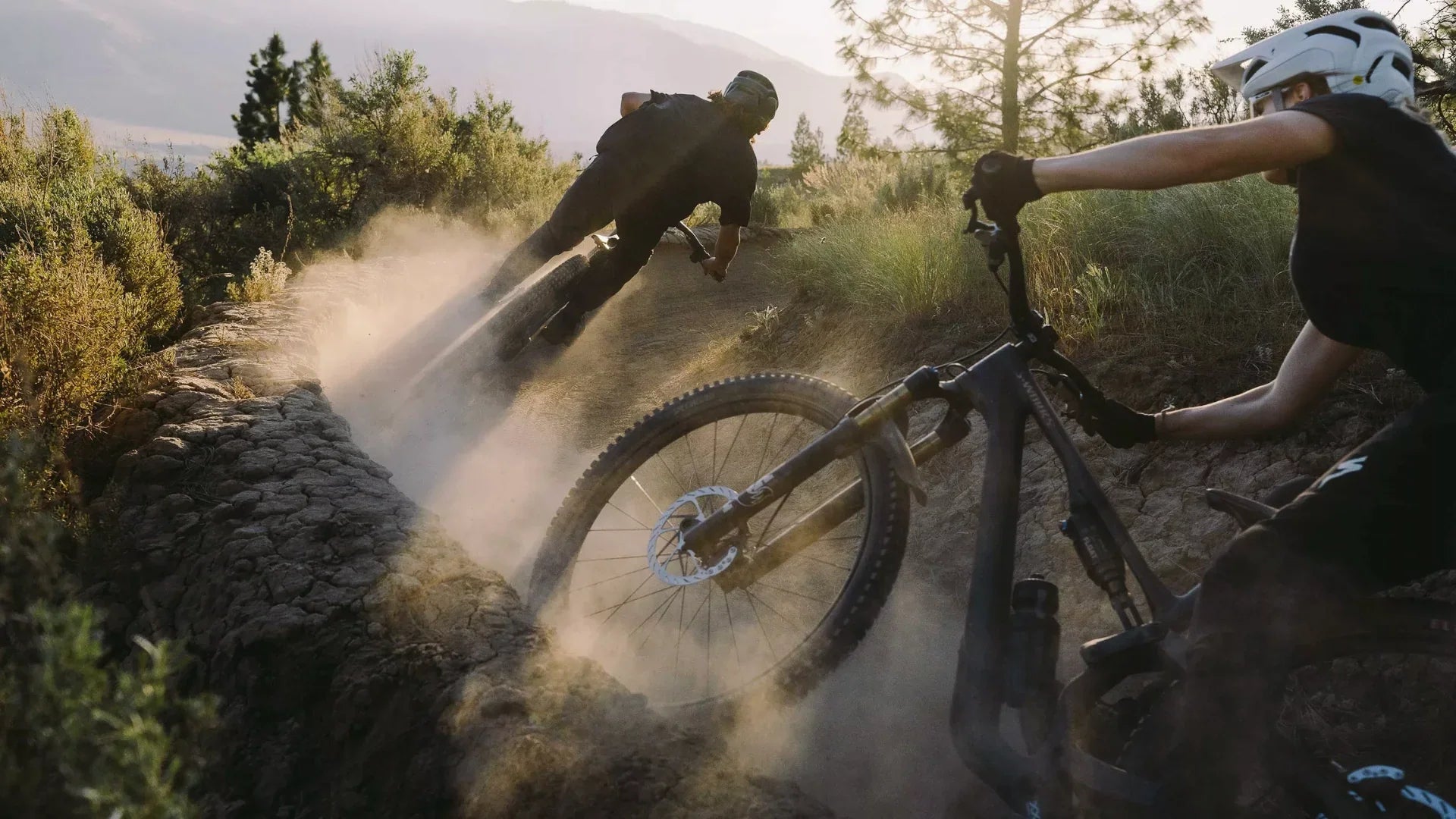 Two mountain bikers wearing helmets ride quickly down a dusty dirt trail surrounded by trees and shrubs, kicking up clouds of dust as they lean into a curve.